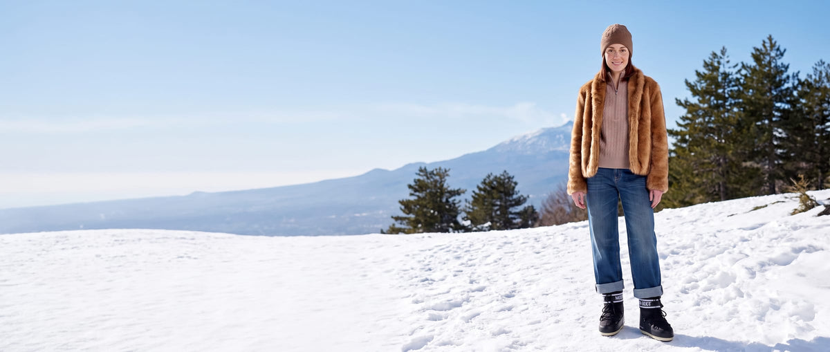 Donna con giacca color miele, cappello e doposci neri sulla neve, con una montagna sullo sfondo