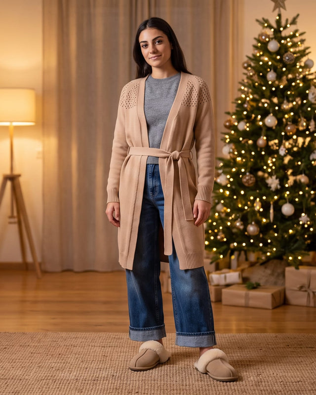 Donna con cardigan beige e pantofole chiuse in salotto davanti a un albero di Natale illuminato